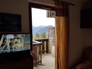 Una habitación con un televisor mostrando un esqueleto, una terraza de madera con barandilla y vistas a montañas.