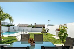 Une table en verre avec un vase de fleurs, un espace de détente extérieur, une piscine et un bâtiment blanc avec un toit en tuiles.