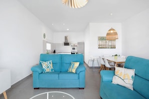 Un salon moderne avec un canapé et des fauteuils, un coin repas avec une table et des chaises, et une cuisine en arrière-plan.