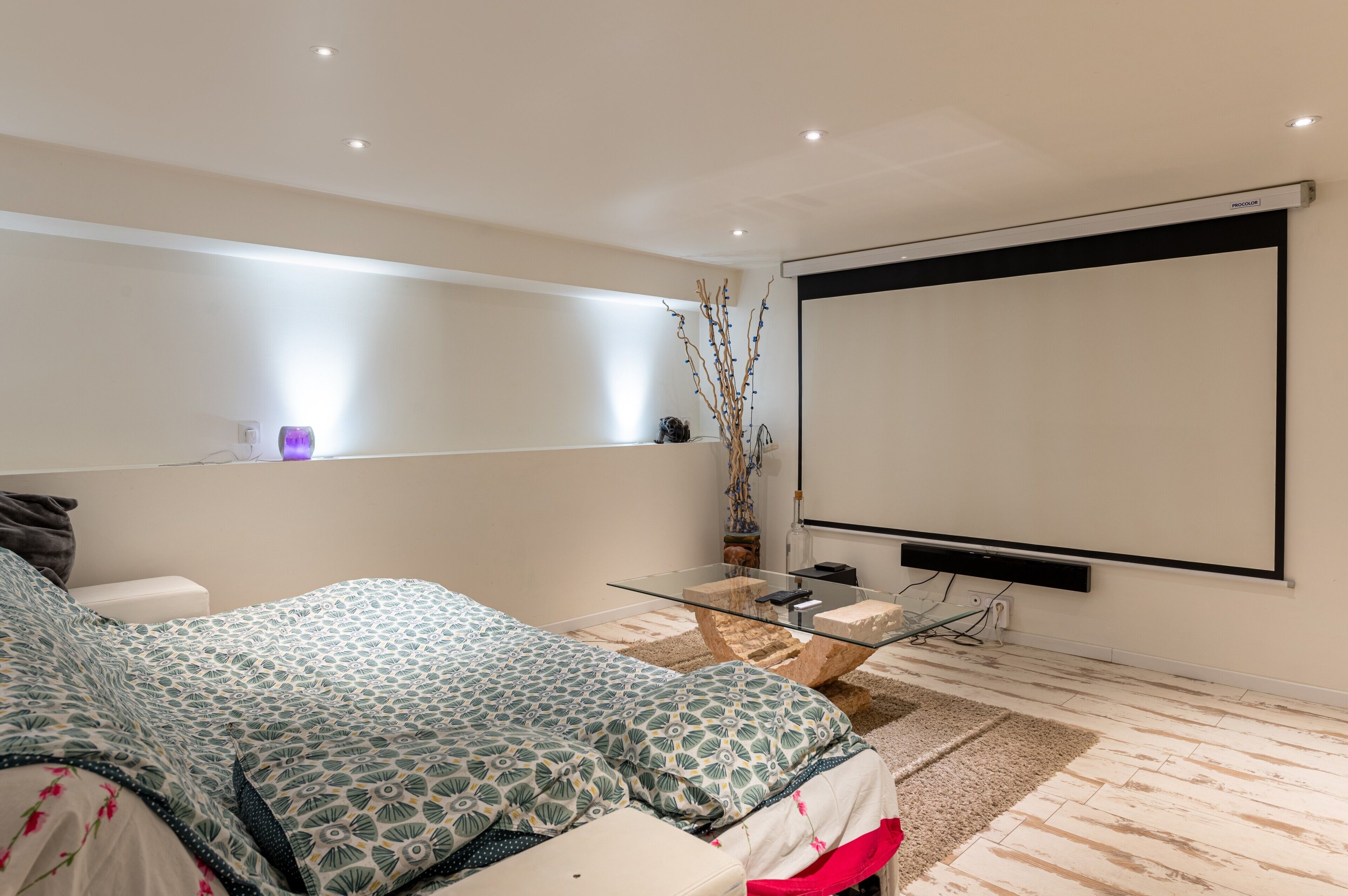 Une chambre moderne comprenant un lit, des tables de chevet, une table basse en verre, un écran de projection et une étagère fixée au mur.