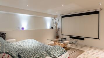 Une chambre moderne comprenant un lit, des tables de chevet, une table basse en verre, un écran de projection et une étagère fixée au mur.