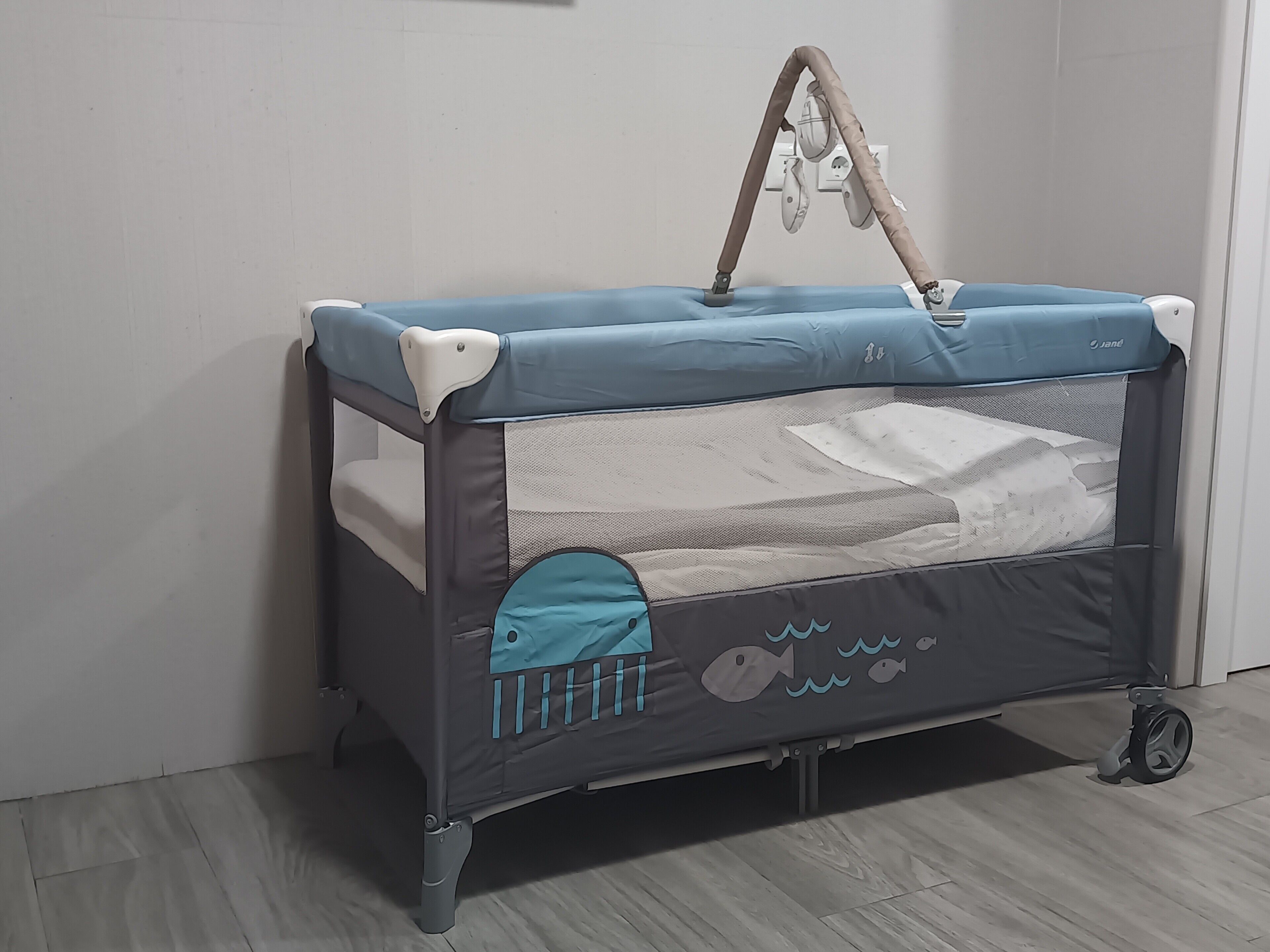 Ein Babybett mit blauer Oberseite, grauen Seiten und einem Netz vorne. Es verfügt über Rollen für den Transport und einen Tragegriff oben.