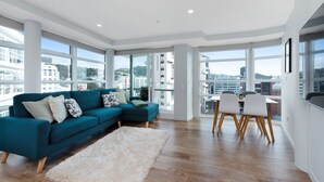 A modern living room with a teal sofa, a white shag rug, and a dining area with wooden chairs. Large windows offer a city view. - Luxury 2 Bedroom at the Pinnacles on Victoria St (Wellington)