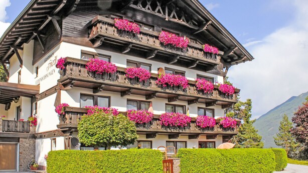 Un hotel de varios pisos con balcones decorados con flores rosadas, un seto cuidado y montañas al fondo.