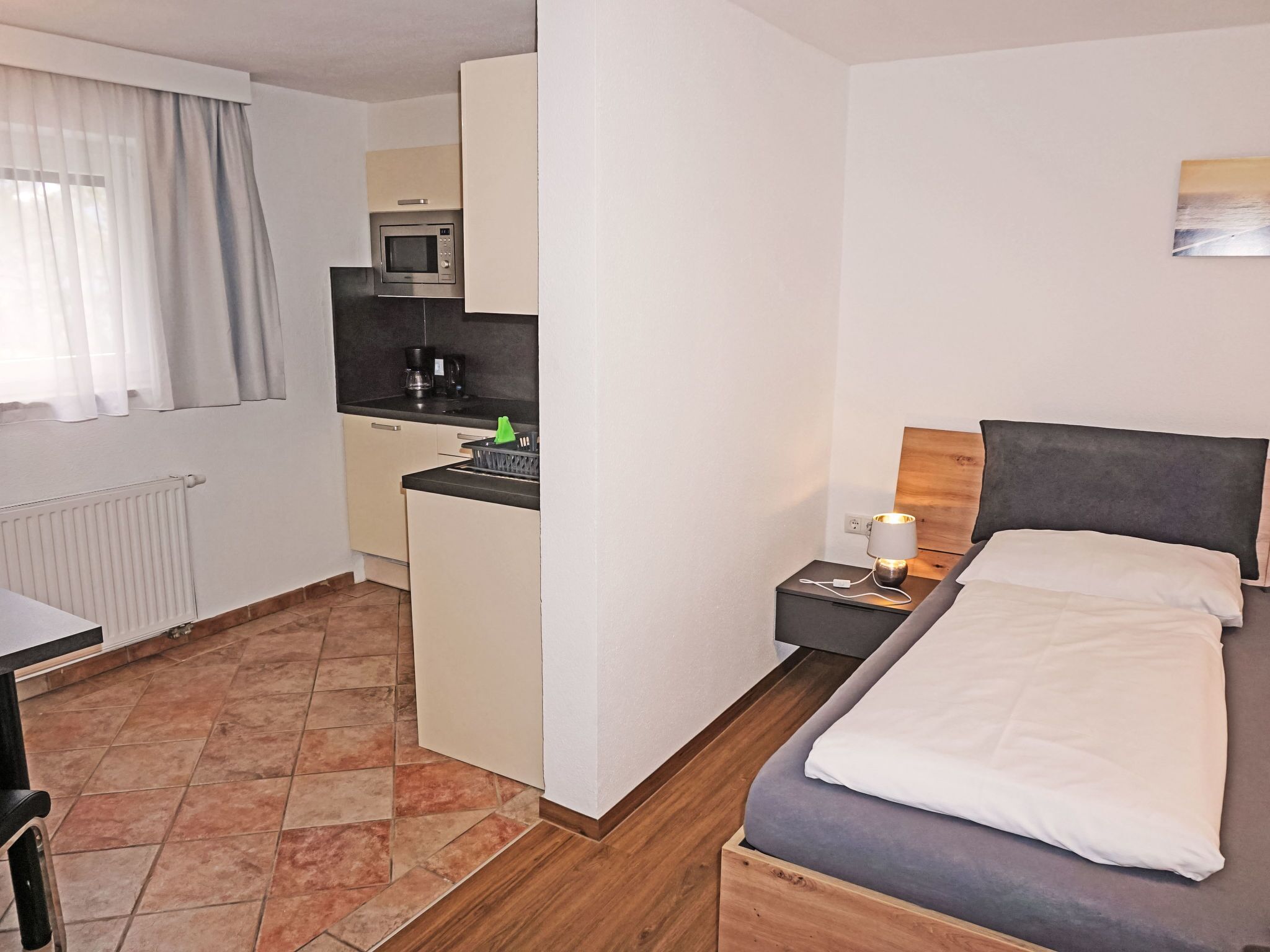 Ein kleines Hotelzimmer mit Küchenzeile, einem Bett, einem Nachttisch mit Lampe und einem fenster mit Vorhängen.