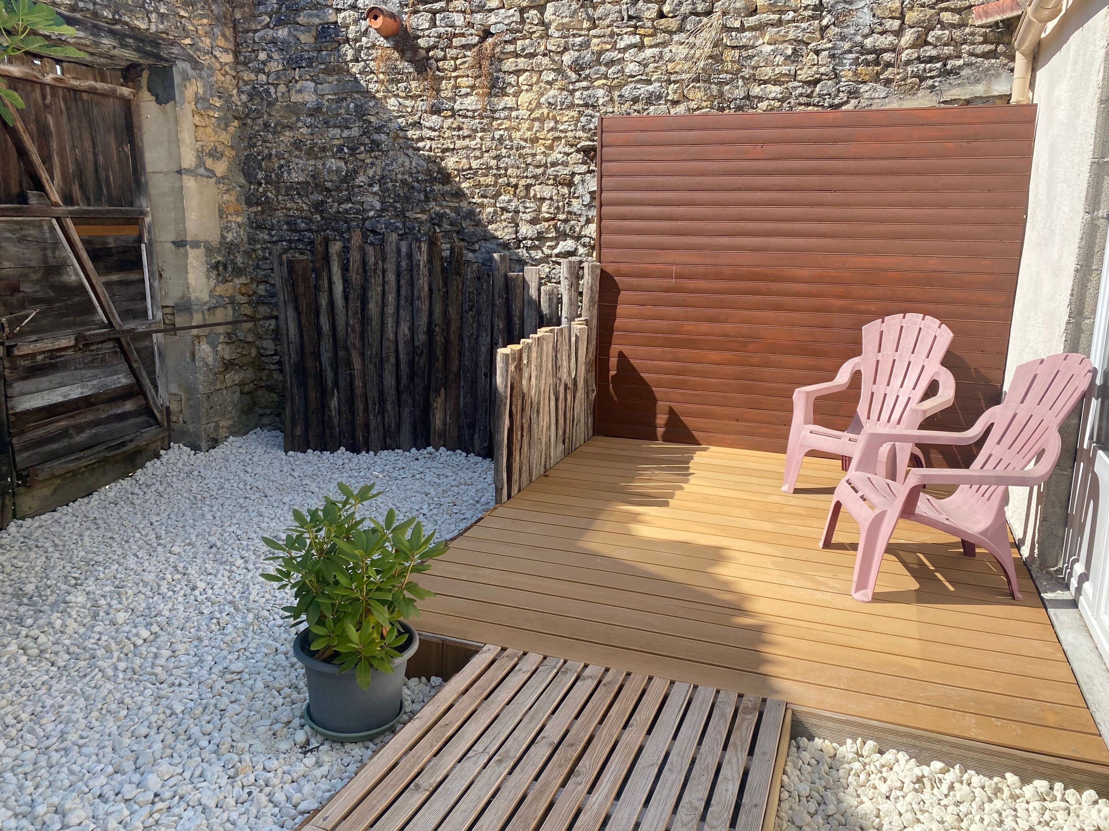 Una terraza de madera con dos sillas rosas, una planta en maceta y un muro de piedra.