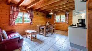 A wooden cabin interior with a sofa, dining table, chairs, a kitchen area, and windows with curtains. - Chalet 'Indépendant 3 Personnes' with Mountain View, Private Terrace and Wi-Fi (Alex)