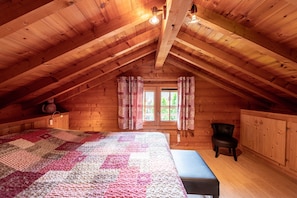 Un dormitorio abrigado en el ático, con techo de madera, cama grande, una silla y una ventana con cortinas.