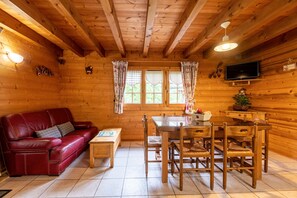 Interior de una cabaña de madera con un sofá de cuero rojo, una mesa de comedor de madera, sillas, un televisor montado en la pared y una ventana con cortinas.