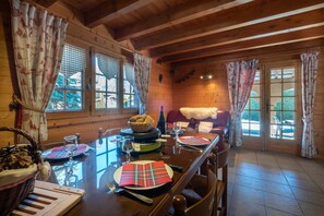 Interior de una cabaña de madera con una mesa de comedor preparada para dos personas, una cesta de pan y una botella de vino.