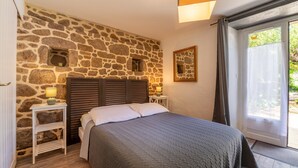 Ein Schlafzimmer mit Steinwand, einem Bett mit grauer Tagesdecke, einem Nachttisch mit Lampe und einem Fenster mit Vorhängen.