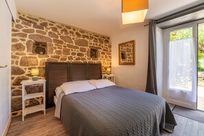 Une chambre avec un mur en pierre, un lit recouvert d’une couette grise, une table de chevet avec une lampe et une fenêtre avec des rideaux.