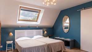 Une chambre avec deux lits, une lucarne, une table de chevet avec une lampe, un banc et un miroir.