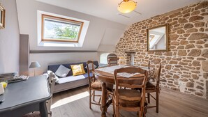 Une pièce chaleureuse avec une table à manger ronde en bois, un canapé avec des coussins, un mur en pierre et une lucarne.