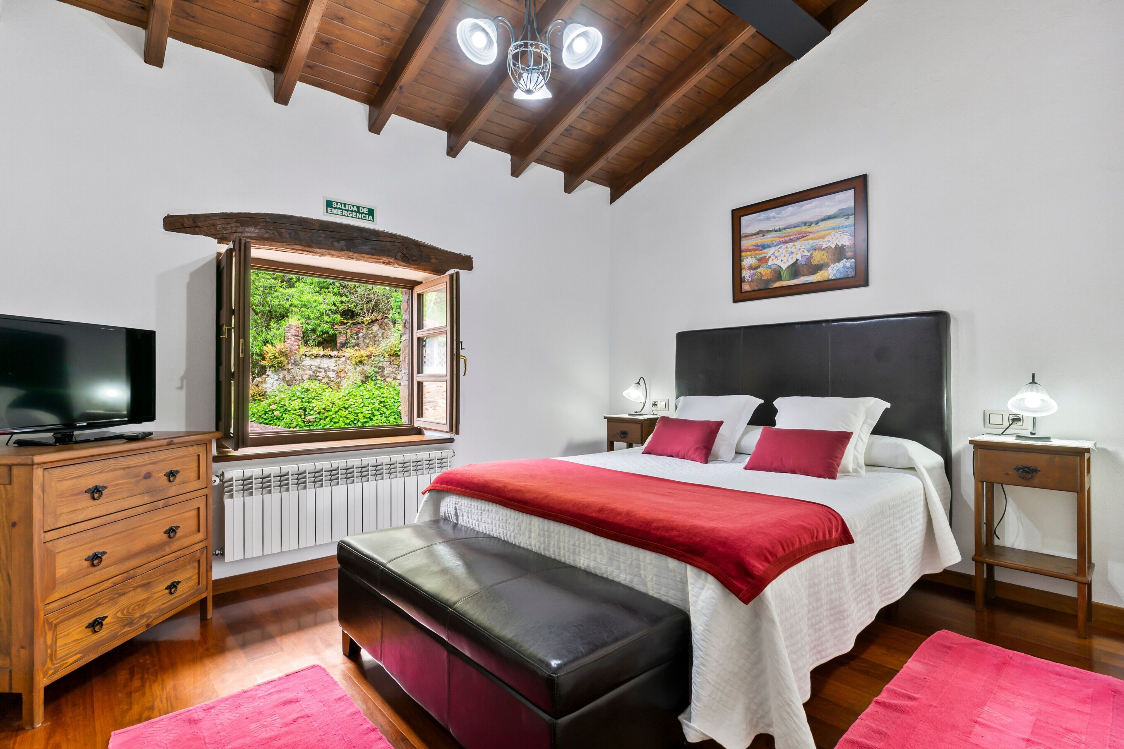 Une chambre avec un grand lit, une télévision, un banc, une fenêtre donnant sur un beau paysage, une table de chevet et un tableau au mur.