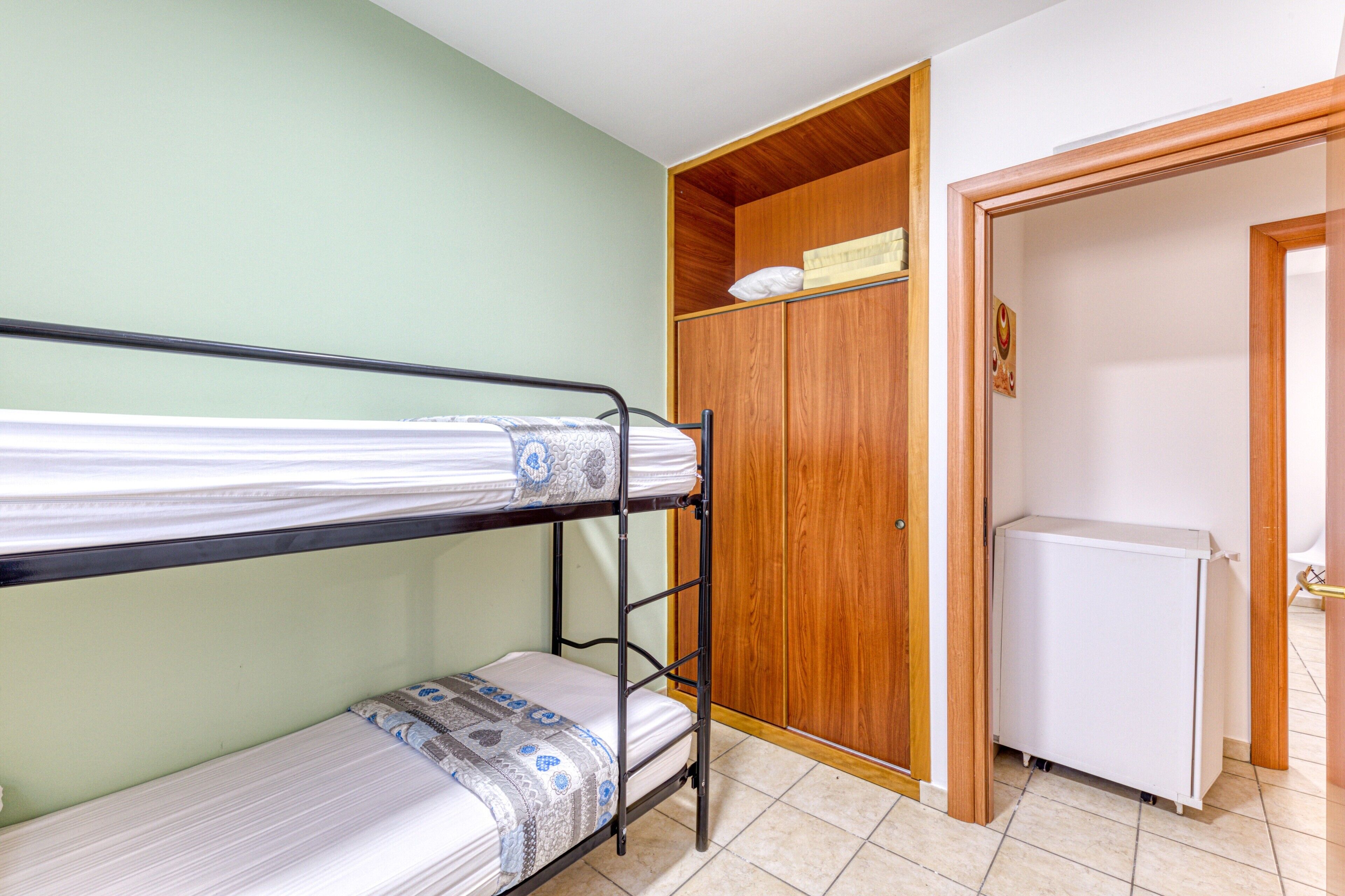 Un letto a castello con copriletto decorato, un armadio in legno e un frigorifero bianco in una stanza dalle pareti verde chiaro.