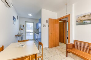 A dining area with a table and chairs, a wooden bench, a kitchen in the background, and a door leading to another room.