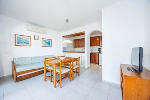 Sala de estar com sofá, mesa de jantar, cadeiras, televisão e cozinha ao fundo.