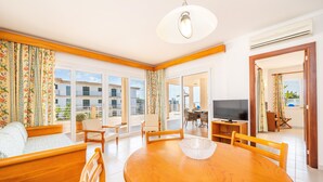 Un salon moderne avec une table à manger en bois, un canapé et des fauteuils, un téléviseur et de grandes fenêtres offrant une vue sur la ville.