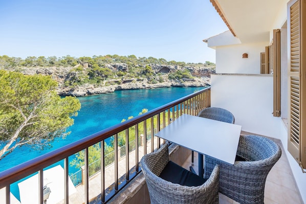 A balcony with wicker furniture, a white table, and a view of the sea and rocky landscape.