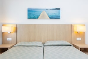 A double bed with a patterned bedspread, two bedside lamps, a wall-mounted power outlet, and a framed picture of a pier extending into a body of water.