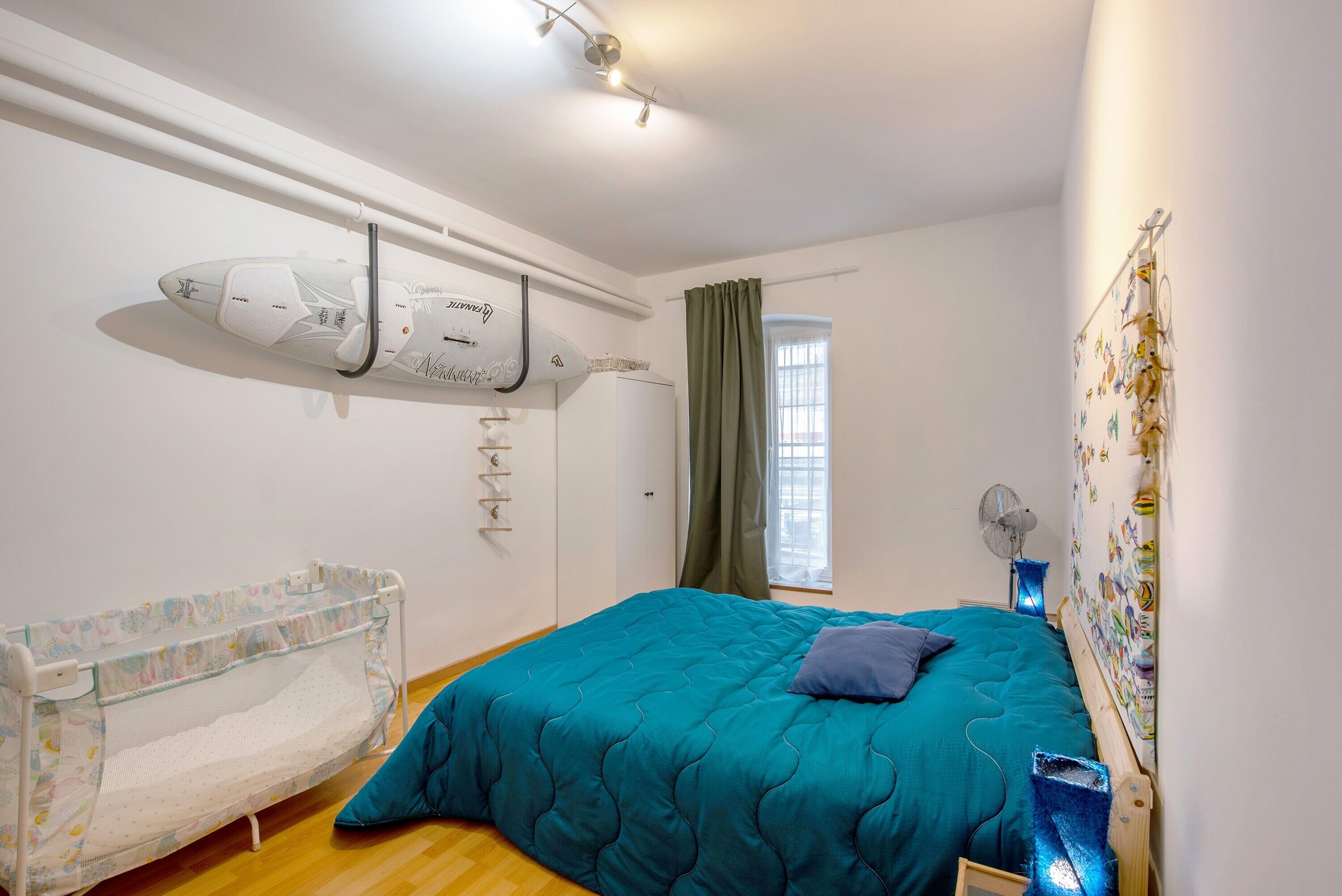 Un dormitorio con cama, cuna, ventana con cortinas y ventilador de pared.