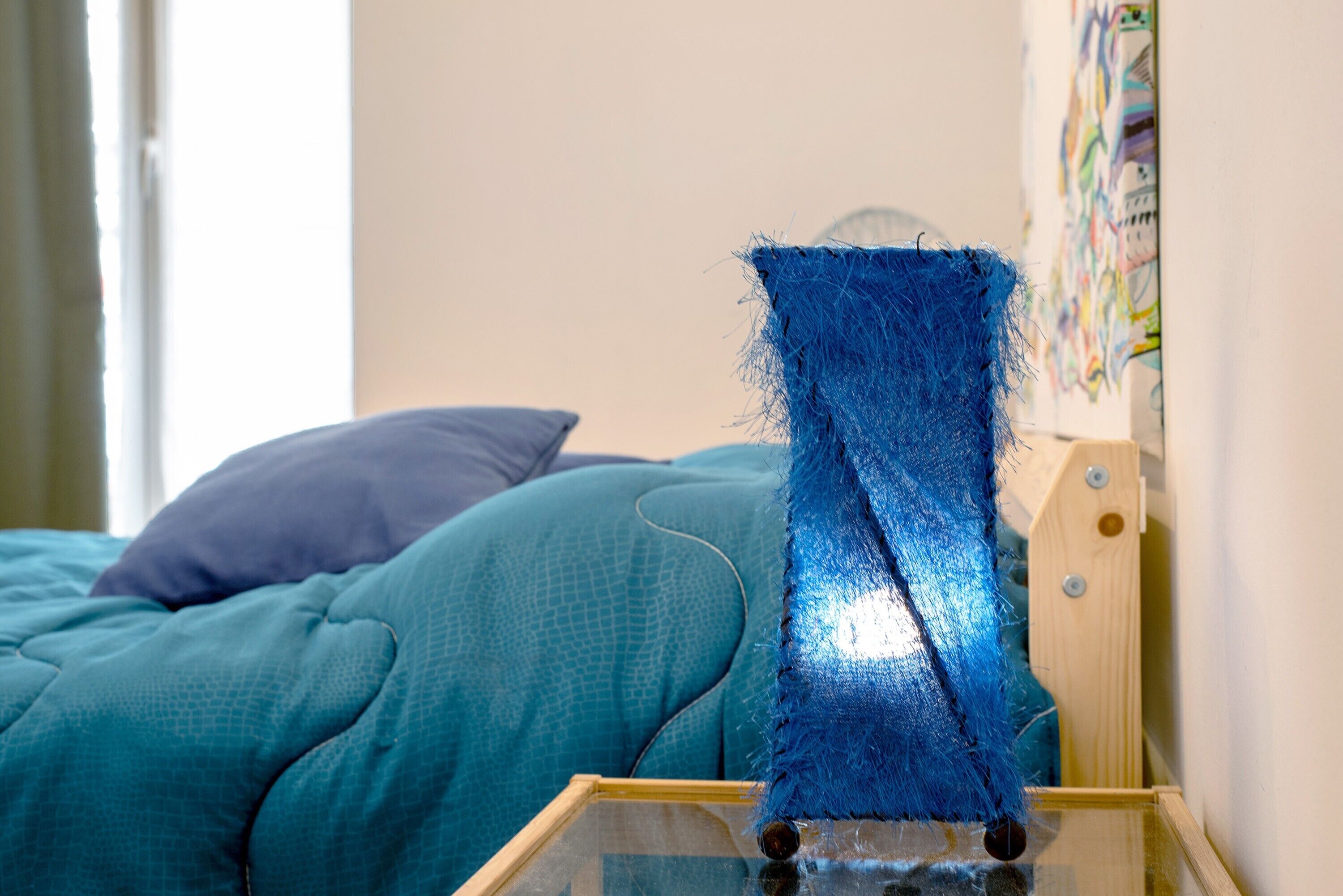 A bed with teal bedding, a blue textured object on a glass table, and a wall with a colorful abstract painting.