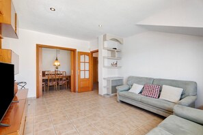 Un salon comprenant un canapé, un téléviseur, un coin repas avec des chaises et une table, ainsi qu’une porte donnant sur une autre pièce.