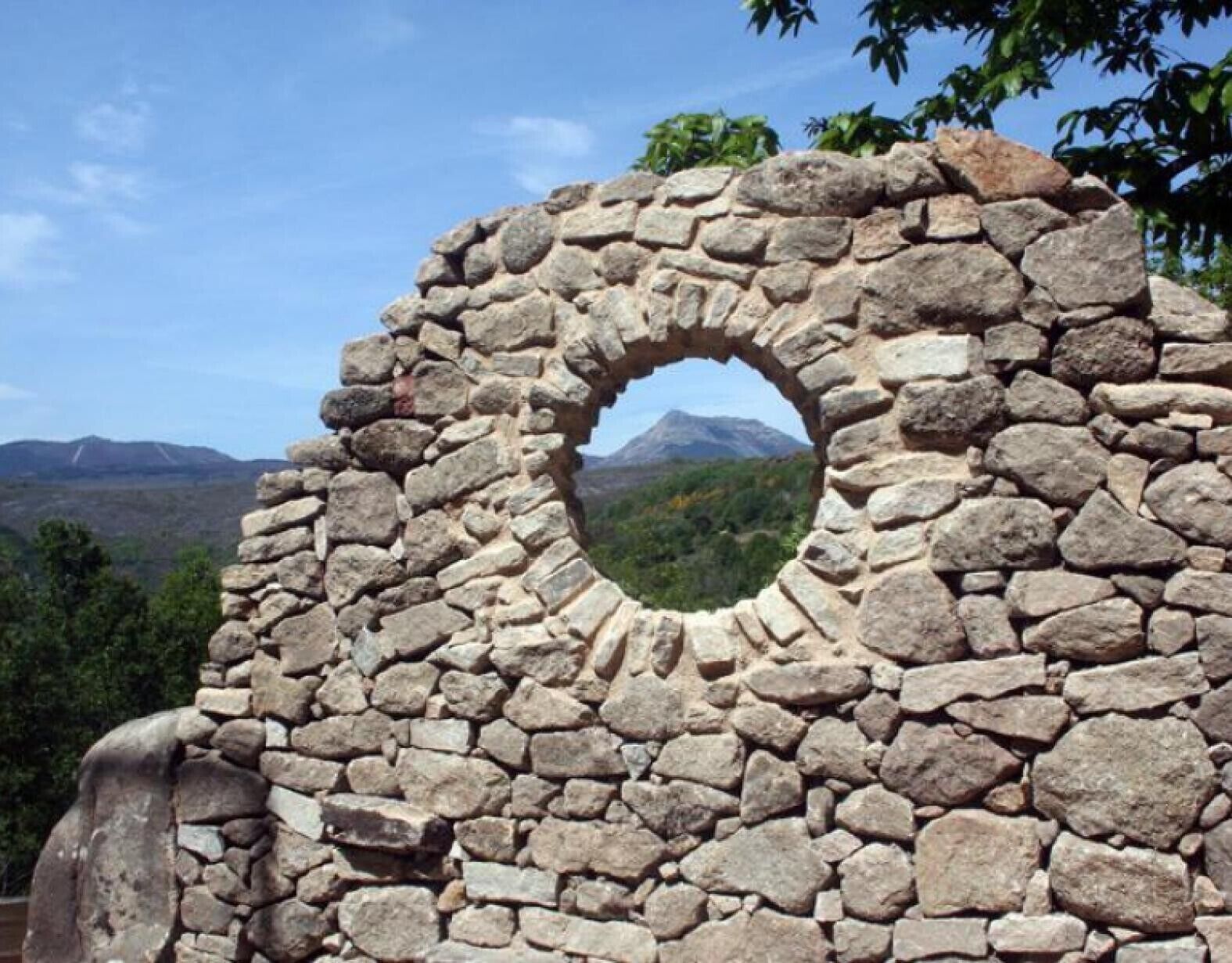 A stone structure with a circular opening, surrounded by greenery and mountains in the background.