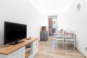 Una sala de estar moderna con una televisión de pantalla plana sobre un mueble de madera, un refrigerador pequeño y una zona de comedor con sillas blancas y una mesa.