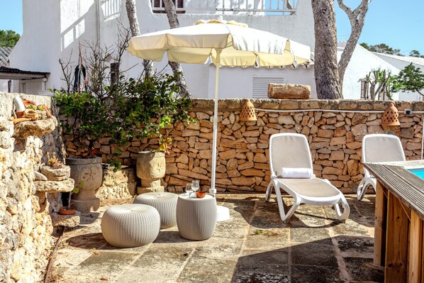 A stone wall, white lounge chairs, a wooden table, and a large umbrella in a courtyard.
