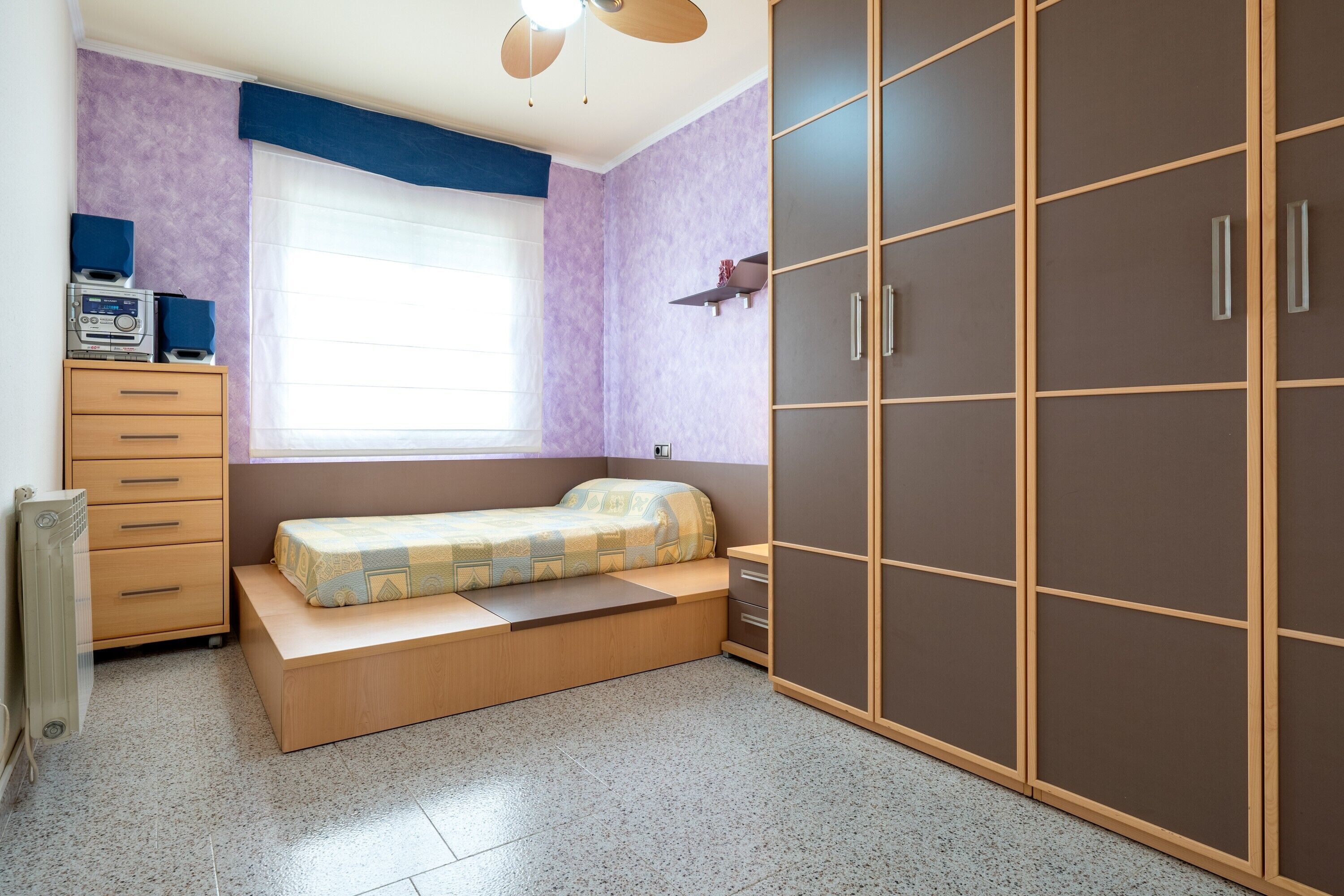 Een slaapkamer met een bed, een kledingkast, een radiator en een raam met zonwering.