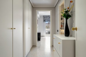 A hallway with white walls, a white door with a brass handle, a white console table with a black vase, and a tiled floor.