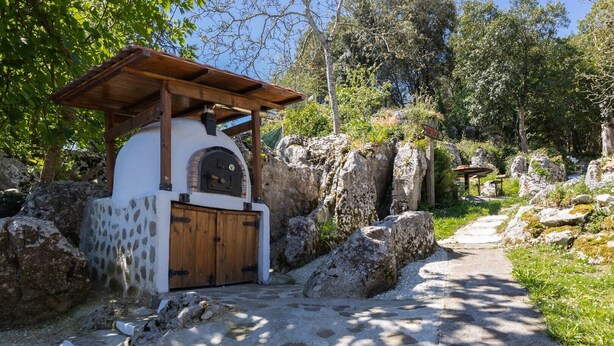 A rustic stone building with a wooden door and a covered entrance, surrounded by large rocks and greenery.