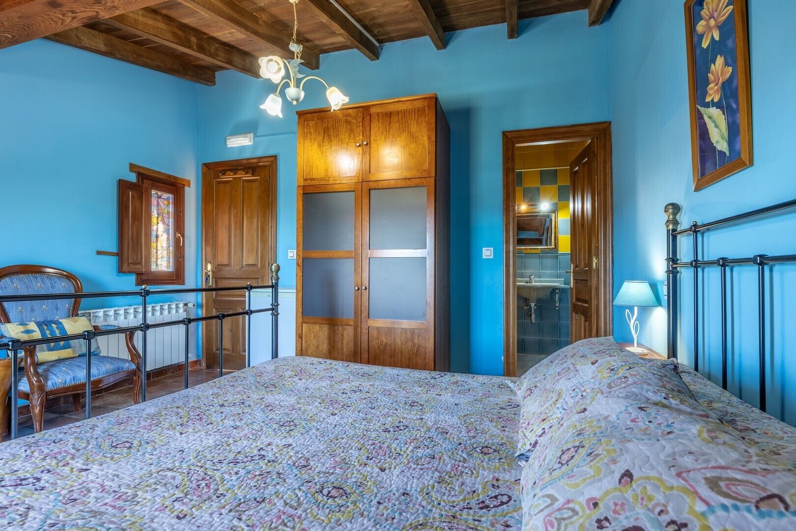 A bedroom with a bed, a chair, a wooden wardrobe, a door, and a ceiling with wooden beams.