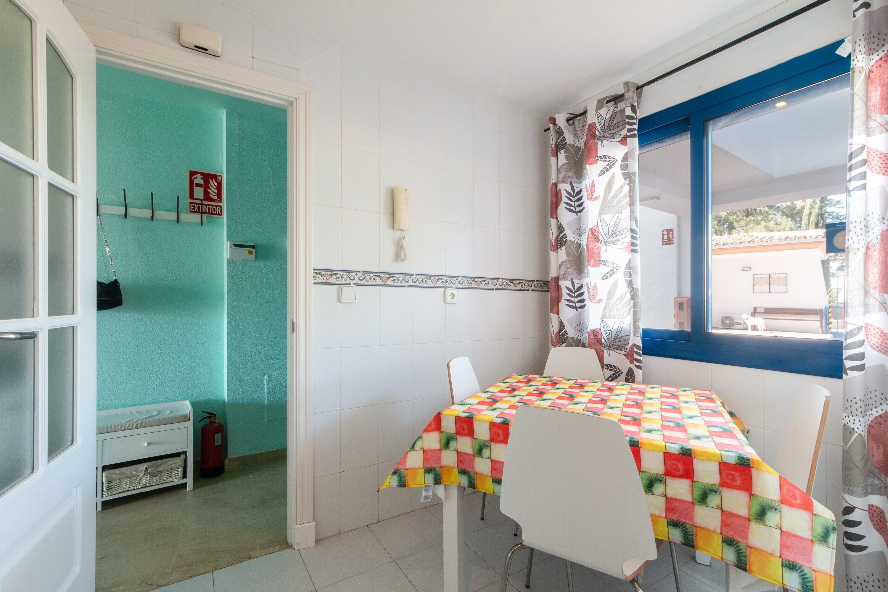 Habitació amb una taula coberta per un mantell de taula colorit, cadires blanques, una finestra amb marc blau i una porta que dona a un bany.