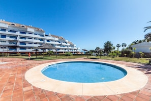 A swimming pool with a tiled deck, a hotel building with blue windows, and palm trees in the background.