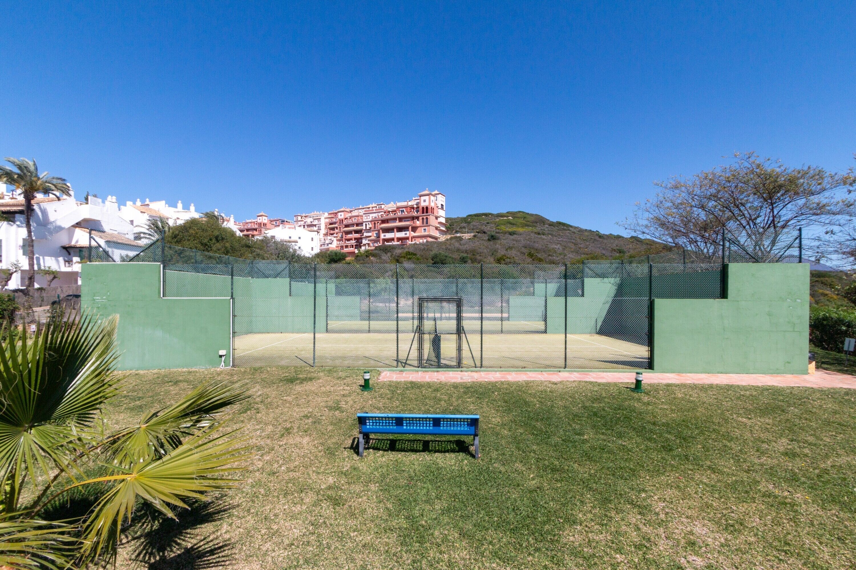 Una cancha de tenis con una valla verde, una banca y edificios al fondo.