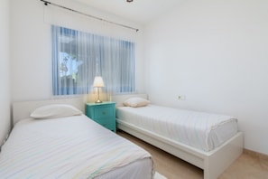 A bedroom with two beds, a nightstand with a lamp, and a window with sheer curtains.