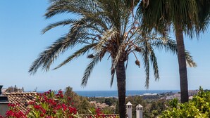 A coastal view with palm trees and red flowers, a clear sky, and a distant cityscape. - Holiday Home 'Townhouse El Naranjal' with Sea View, Wi-Fi and Air Conditioning (Marbella)