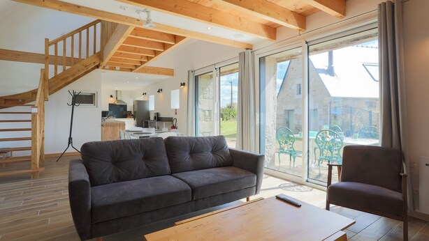 A modern living room with a sofa, a wooden coffee table, and a chair. There is a staircase with wooden beams and a large sliding glass door leading to an outdoor area. - Holiday Home 'Gite Des Hauts Grève - Near the Sea' with Terrace, Garden & Wi-Fi (Saint-Michel-en-Grève)