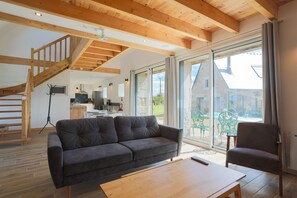 A modern living room with a sofa, a wooden coffee table, and a chair. There is a staircase with wooden beams and a large sliding glass door leading to an outdoor area.