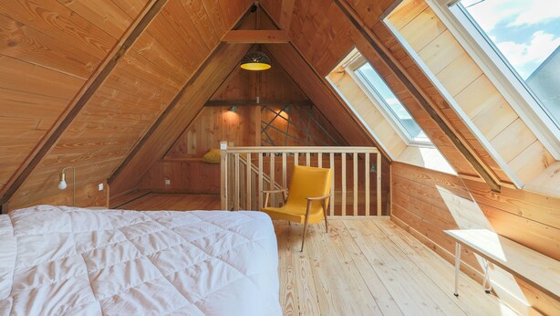 Une chambre à coucher aménagée dans un espace sous les toits, avec un plafond en pente, des murs en bois, un lit, une chaise jaune, un banc blanc et des fenêtres de toit.