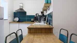 Une cuisine compacte avec un plan de travail en bois, une hotte de cuisine en acier inoxydable et un évier. On y trouve des chaises bleues et une étagère avec des tasses turquoise.