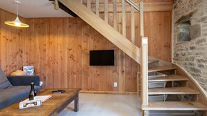 Un intérieur en bois avec un escalier, un téléviseur à écran plat, un canapé et une table basse sur laquelle se trouvent une bouteille et des télécommandes.