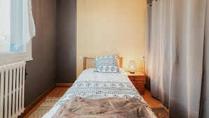 A small bedroom with a bed, a nightstand, a lamp, a radiator, and a window with curtains.