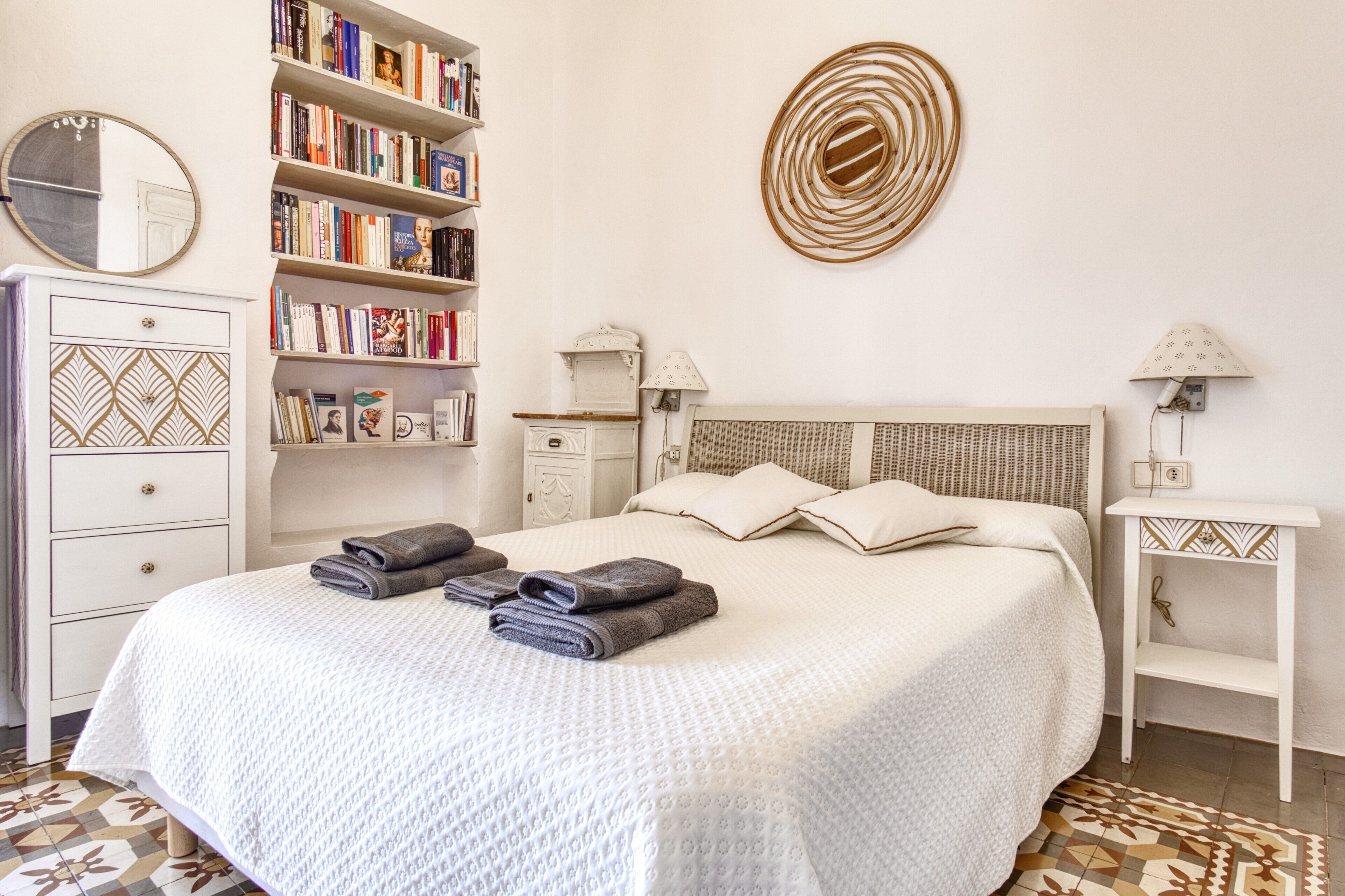 A neatly arranged bedroom with a large bed, a bookshelf, a mirror, and a patterned rug.