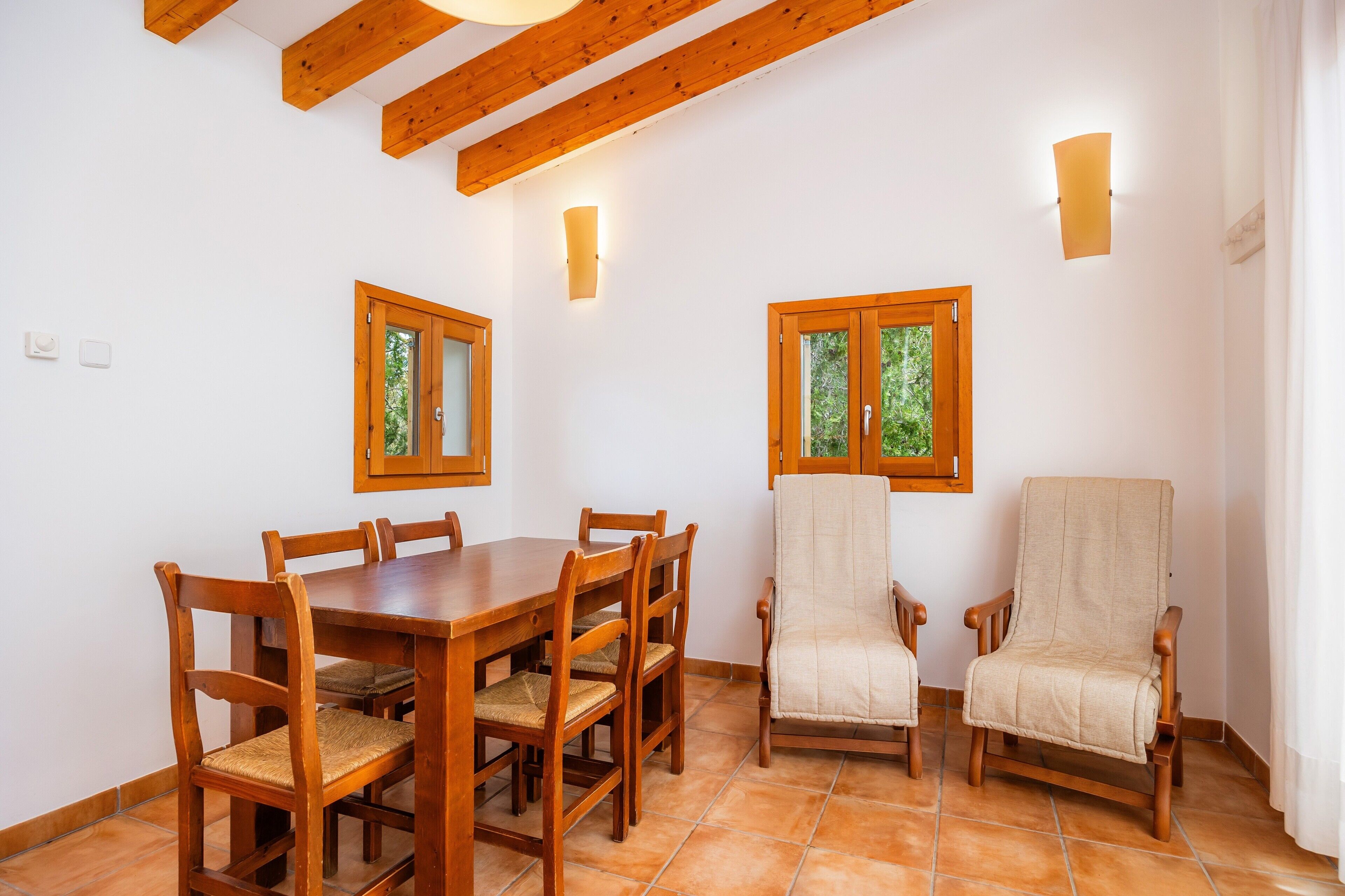 Un comedor con una mesa y sillas de madera, dos lámparas fijas en la pared y ventanas con marcos de madera.