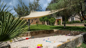 Zona de juegos arenosa con toldo, rodeada de vegetación y un muro de piedra.