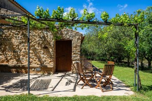 Un edificio de piedra con puerta de madera, mesas al aire libre con sillas de madera, y una pérgola cubierta de hiedra.